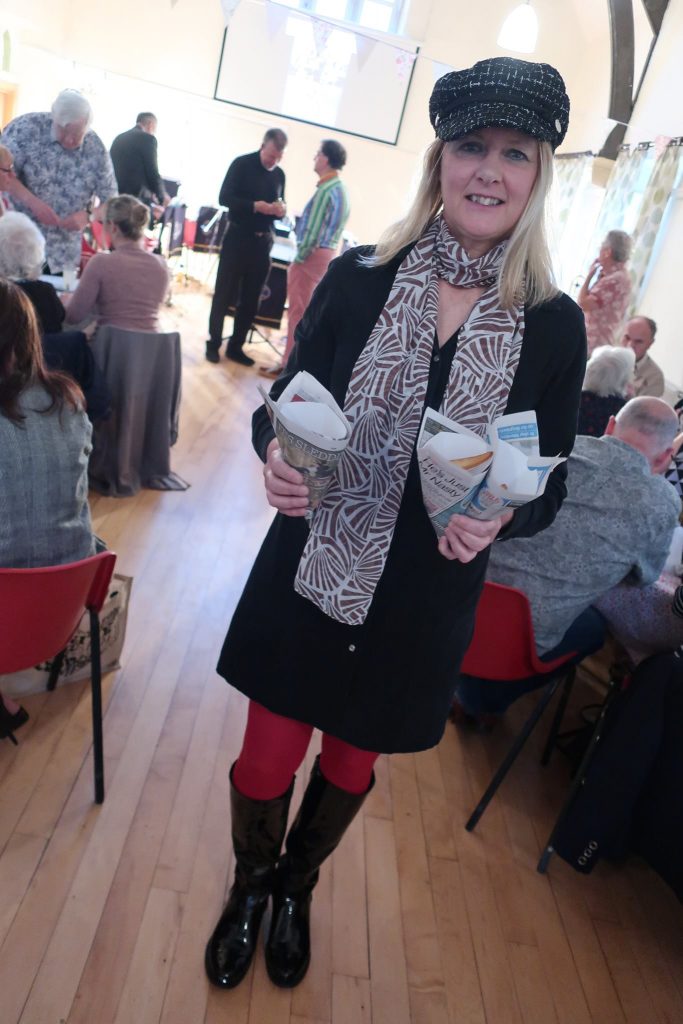 Rachel is wearing a sixties outfit including a black hat and boots and red tights. She is holding 3 newspaper cones of chips.