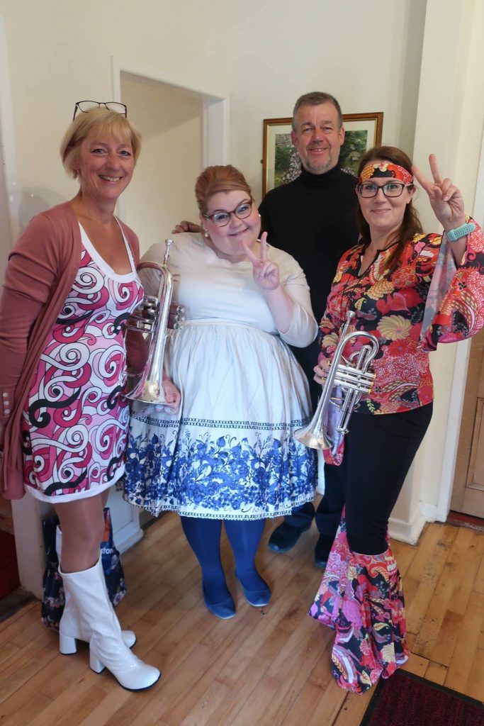 Four band members, some holding their instruments, in sixties outfits, doing peace signs.